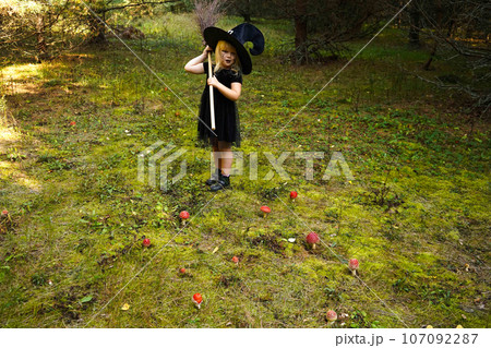 A girl in a witch costume stands with a broom, in an autumn forest. Halloween mysterious lady in woods 107092287