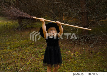 A girl in a witch costume stands with a broom, in an autumn forest. Halloween mysterious lady in woods 107092309