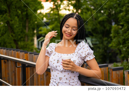 African young attractive woman in glasses stands on a bridge with a paper cup of coffee on the street during the day in the park in summer African young attractive woman in glasses stands on a bridge with a paper cup of coffee on the street during the day in the park in summer 107093472