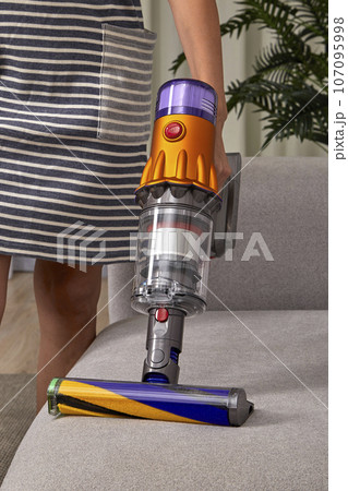 Beautiful slender caucasian girl in a striped dress is vacuuming the sofa upholstery with a modern wireless vertical vacuum cleaner with a special nozzle for fabric. 107095998