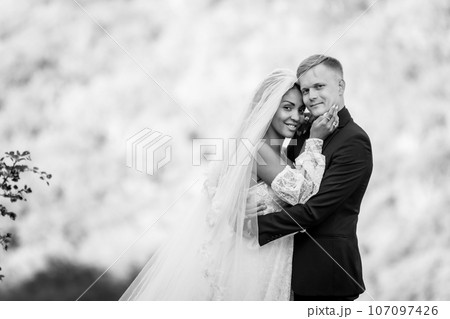Happy newlyweds against the backdrop of evening sunny foliage, the couple looks into the frame, black and white version 107097426
