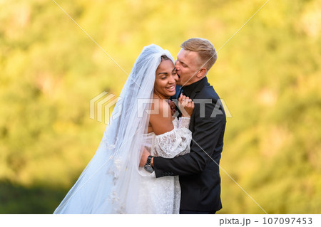 Happy newlyweds are hugging against the backdrop of sunny evening foliage, the guy is trying to cheerfully bite the girl Happy newlyweds are hugging against the backdrop of sunny evening foliage, the guy is trying to cheerfully bite the girl 107097453
