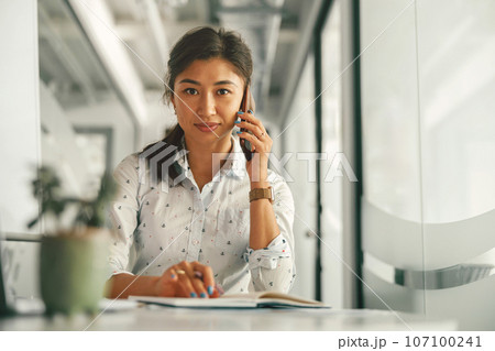Businesswoman talking phone with client and making notes while sitting in office. High quality photo 107100241