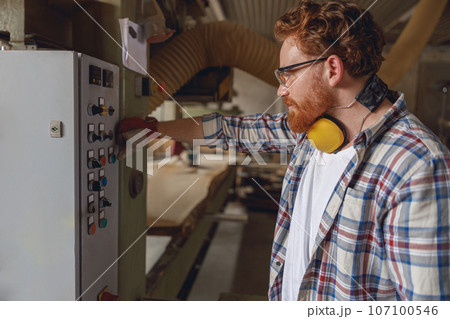Professional carpenter in furniture factory pressing button on machine to start production 107100546