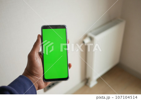 holding smart phone with green screen against White radiator on grey white wall holding smart phone with green screen against White radiator on grey white wall 107104514