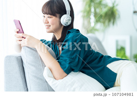 スマホを操作しながらリビングのソファで音楽を聴く若い女性 107104981