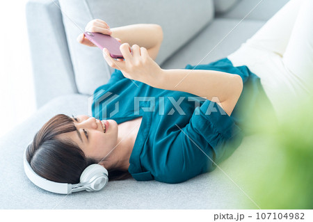 スマホを操作しながらリビングのソファで音楽を聴く若い女性 107104982