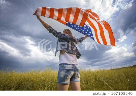 Beautiful young girl holding an American flag on the wind in a rapeseed field. Summer landscape against the blue sky. 107108340