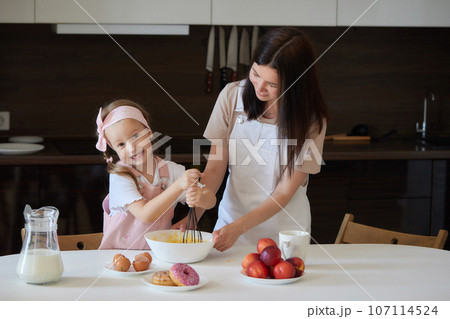 Mother and daughter preparing a sweet cake using flour, milk, sitting on chairs at a table in a modern kitchen. Girl holding a whisk, stirring eggs in a bowl, preparing pancake dough with her mom. Mother and daughter preparing a sweet cake using flour, milk, sitting on chairs at a table in a modern kitchen. Girl holding a whisk, stirring eggs in a bowl, preparing pancake dough with her mom. 107114524
