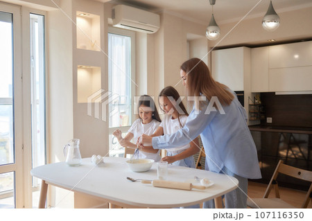 Cute little girl and her beautiful mom in aprons are playing and laughing while kneading the dough in the kitchen. 107116350