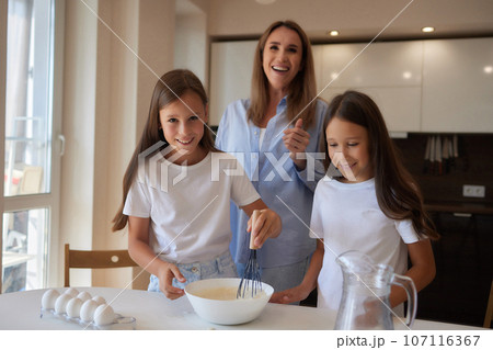 Cute little girl and her beautiful mom in aprons are playing and laughing while kneading the dough in the kitchen. 107116367