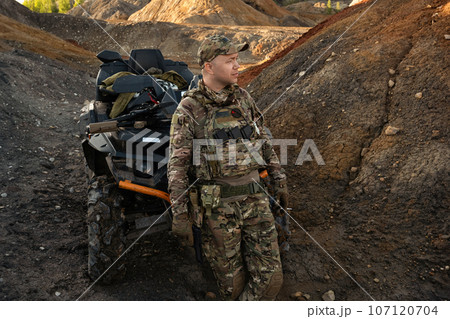 a man in a military uniform with an ATV and a machine gun in his hands is standing in the mountains. a man in a military uniform with an ATV and a machine gun in his hands is standing in the mountains. 107120704