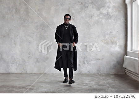 Stylish man walking near grey textured studio wall, wearing black coat, turtleneck, pants, loafers shoes. Fashion african american male model in Fashionable clothes, total black Stylish man walking near grey textured studio wall, wearing black coat, turtleneck, pants, loafers shoes. Fashion african american male model in Fashionable clothes, total black 107122846