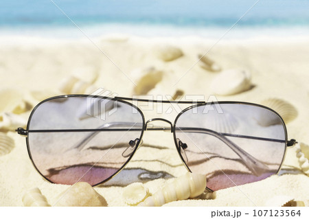 Purple and pink gradient color sunglasses on beach. Plenty of seashells on tropical sand. Sea view. Purple and pink gradient color sunglasses on beach. Plenty of seashells on tropical sand. Sea view. 107123564
