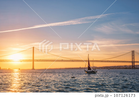 View of 25 de Abril Bridge famous tourist landmark of Lisbon connecting Lisboa and Almada over Tagus river with tourist yacht silhouette at sunset. Lisbon, Portugal 107126173