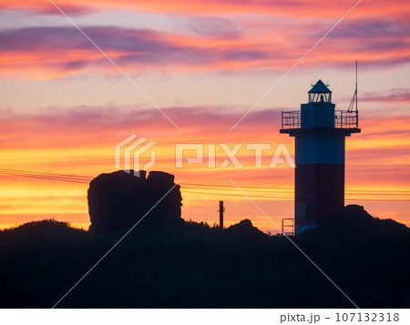 北海道稚内市沖の日本海に浮かぶ利尻島の沓形岬公園の夕焼けを背景とした赤白の灯台と岩のシルエット 北海道稚内市沖の日本海に浮かぶ利尻島の沓形岬公園の夕焼けを背景とした赤白の灯台と岩のシルエット 107132318
