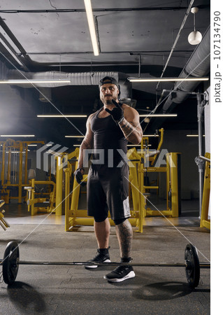 Attractive, tattooed, bearded man in black sport gloves, shorts, vest, cap and sneakers. Showing cool sign, barbell is on floor. Dark gym. Full length Attractive, tattooed, bearded man in black sport gloves, shorts, vest, cap and sneakers. Showing cool sign, barbell is on floor. Dark gym. Full length 107134790