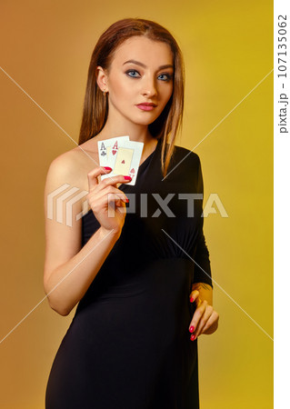 Blonde model with bright make-up, in black dress is showing two aces, posing against colorful background. Gambling, poker, casino. Close-up. 107135062