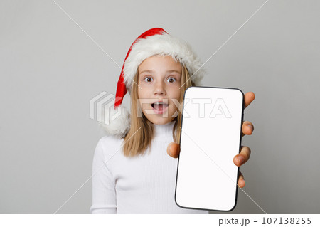 Shocked kid girl in Santa hat holds smartphone with blank empty white screen display 107138255