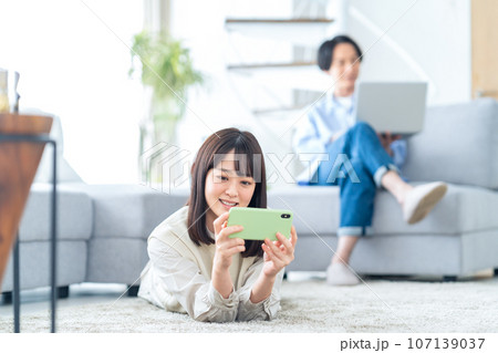 スマホを見ながらリビングでくつろぐ仲良しな若いカップル スマホを見ながらリビングでくつろぐ仲良しな若いカップル 107139037