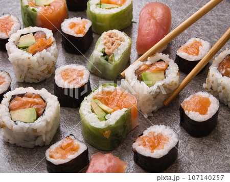 on a table, assorted rolls, with salmon, tuna and cucumber, laid out in a row, wooden sticks take one 107140257