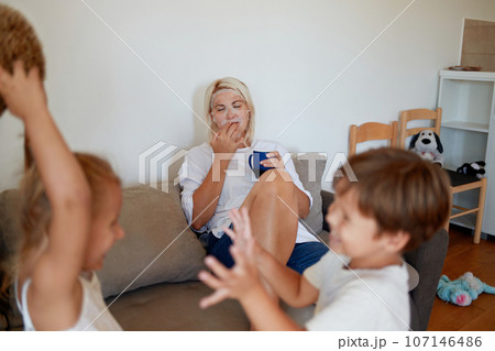 A mom is applying fabric cosmetic mask on the couch while her two children are playing. 107146486