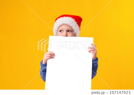 Cute little girl in Santa Claus hat with a white blank paper sheet. Christmas, x-mas, New Year, Winter, people, advertisement, sale concept. Cute little girl in Santa Claus hat with a white blank paper sheet. Christmas, x-mas, New Year, Winter, people, advertisement, sale concept. 107147023