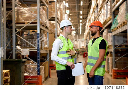 Two warehouse managers in work wear organizing distribution in warehouse storage area. 107148851
