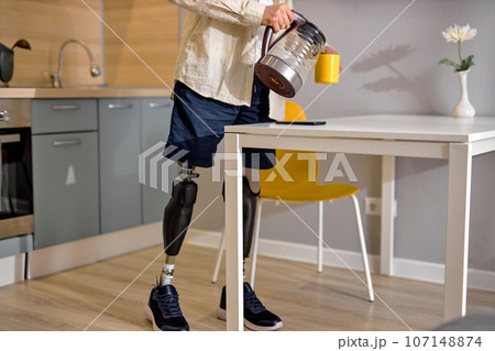 disabled guy with leg prosthesis in kitchen at home, making tea in the morning disabled guy with leg prosthesis in kitchen at home, making tea in the morning 107148874