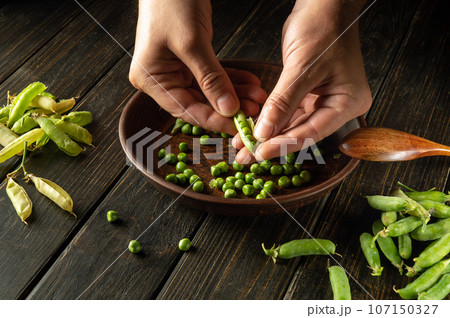 Close-up of the cook hands on the kitchen table peeling green pea pods. Organic peasant food after harvest Close-up of the cook hands on the kitchen table peeling green pea pods. Organic peasant food after harvest 107150327