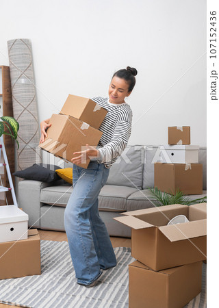 Welcome Home: A happy woman, moving into her new home, enters the living room with a pile of cardboard moving boxes, welcoming the sense of belonging Welcome Home: A happy woman, moving into her new home, enters the living room with a pile of cardboard moving boxes, welcoming the sense of belonging 107152436