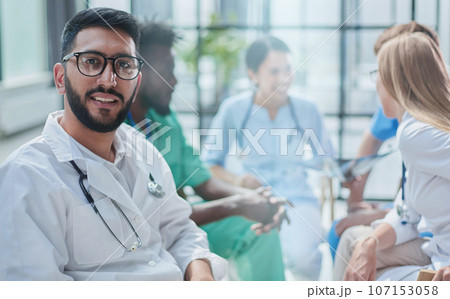 a young doctor in glasses looks at the camera against the background of his colleagues' discus a young doctor in glasses looks at the camera against the background of his colleagues' discus 107153058