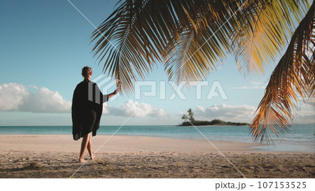 Woman walking barefoot white sand beach at sunset. Tourist girl in black dress enjoy tropical island landscape, blue ocean water and coconut palms. Travel, tourism, holiday. Back view Woman walking barefoot white sand beach at sunset. Tourist girl in black dress enjoy tropical island landscape, blue ocean water and coconut palms. Travel, tourism, holiday. Back view 107153525