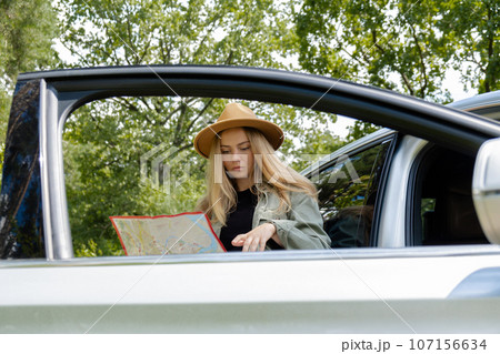 Smiling young woman in hat stoped on road to check the route on travel map. Local solo travel on weekends concept. Exited woman explore freedom outdoors in forest. Unity with nature lifestyle, rest 107156634