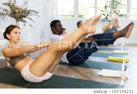 Multinational women and men practicing upward boat pose of yoga on black mat in fitness room Multinational women and men practicing upward boat pose of yoga on black mat in fitness room 107158231