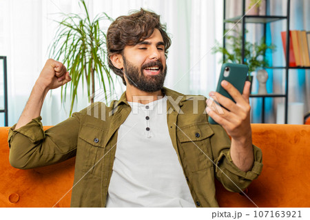 Middle eastern young man at home use smartphone celebrating success victory winning play online game Middle eastern young man at home use smartphone celebrating success victory winning play online game 107163921