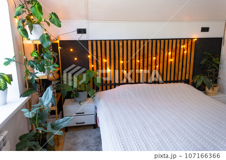 Loft style bedroom interior, black wall with wooden slats, metal bed, retro light bulbs garland, potted plants on a trapezoidal window in the attic. Modern Green House 107166366