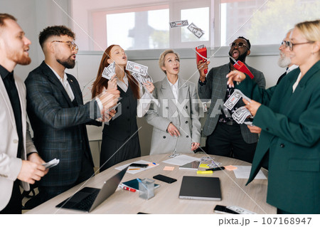 Cheerful business team of mixed-aged diverse coworkers having fun, jumping, rejoicing victory. Happy excited African-American male boss in sunglasses and suit using money gun shooting with banknotes. 107168947