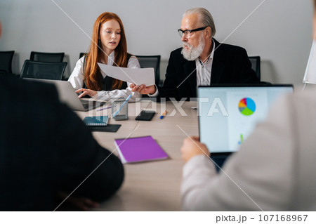 Portrait of mature adult male boss team leader giving paper report business plan to employees group at corporate briefing table. Corporate meeting office workers in modern coworking space. 107168967