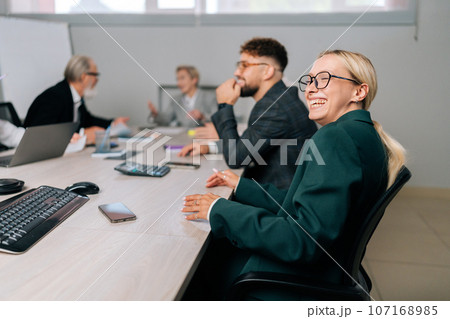 Pretty businesswoman in suit laughing at funny joke with diverse-ages multiethnic coworkers in office. Friendly work team enjoying positive emotions together. Happy colleagues staff group having fun 107168985