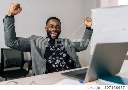 Portrait of successful African businessman in suit looking excited by good news online. Lucky winner man sitting at office desk raising hand in yes gesture celebrating business success win result. 107168987