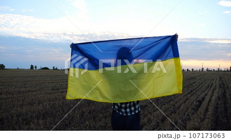Ukrainian lady walking with national blue-yellow banner on barley meadow at sunrise. Woman going with raised flag of Ukraine above her head on wheat field at sunset. Victory against rusian aggression. Ukrainian lady walking with national blue-yellow banner on barley meadow at sunrise. Woman going with raised flag of Ukraine above her head on wheat field at sunset. Victory against rusian aggression. 107173063
