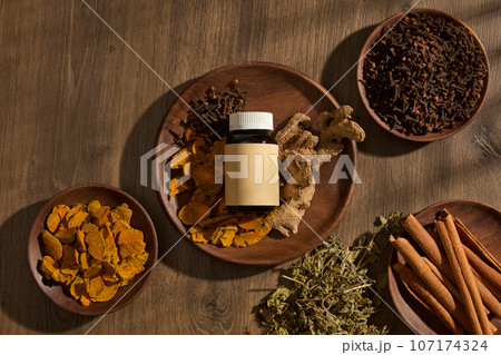 On wooden plates containing folk medicines such as ginger, turmeric, cloves, cinnamon and mint, an empty label medicine bottle is displayed. Wooden background for advertising with traditional ideas On wooden plates containing folk medicines such as ginger, turmeric, cloves, cinnamon and mint, an empty label medicine bottle is displayed. Wooden background for advertising with traditional ideas 107174324