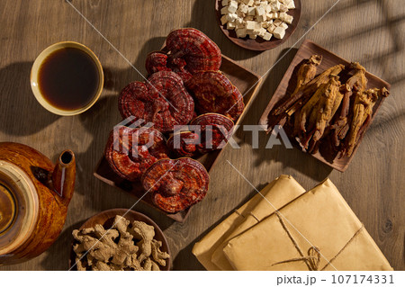 The scene advertises rare natural herbs that are often used to make health supplements. Ganoderma, red ginseng roots, ginger and poria cocos are stored on wooden trays, decorated on wooden background 107174331