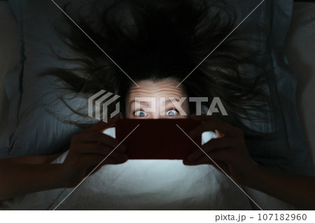 Amazed face of young woman staring up at her smartphone late at night in bed. 107182960