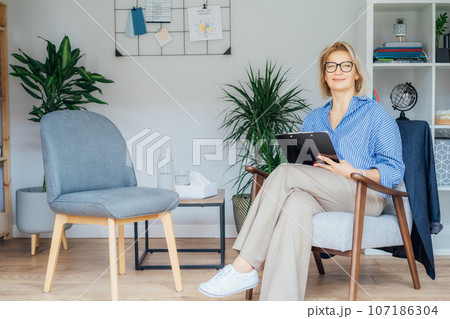 Professional Psychotherapy. Successful female Psychologist Smiling To Camera Sitting On armchair In Office. Mature 50s middle-age professional portrait of teacher, coach, mentor, therapist, counselor Professional Psychotherapy. Successful female Psychologist Smiling To Camera Sitting On armchair In Office. Mature 50s middle-age professional portrait of teacher, coach, mentor, therapist, counselor 107186304