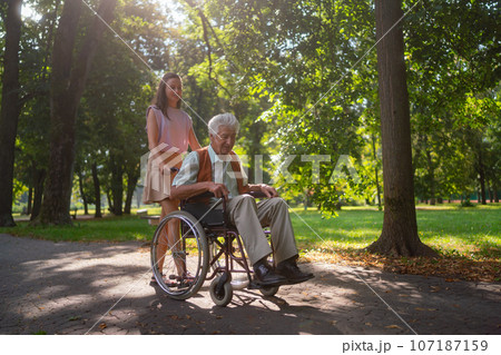 Granddaughter pushing the grandfather in wheelchair in city park. 107187159