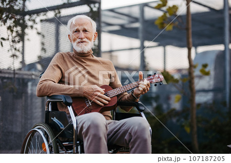 Senior man sitting in a wheelchair outdoors, playing the ukulele. Senior man sitting in a wheelchair outdoors, playing the ukulele. 107187205
