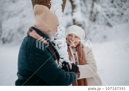 Elegant senior couple standing by a tree in the snowy park, during cold winter snowy day. Elegant senior couple standing by a tree in the snowy park, during cold winter snowy day. 107187391