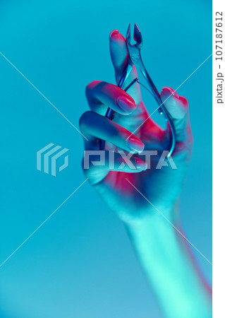 Female hand with nude, natural nails, manicure holding nippers isolated over blue background in neon light 107187612